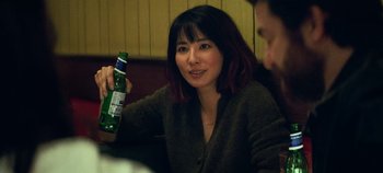 Movie still from “Heart of Stone” (2023), directed by Tom Harper – A woman sitting at a table holding a bottle of beer; Close Up shot, Over the shoulder angle