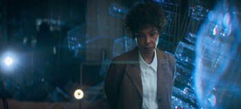 Movie still from “Heart of Stone” (2023), directed by Tom Harper – A woman in a brown jacket standing next to a glass wall; Medium shot, Over the shoulder angle