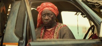 Movie still from “Heart of Stone” (2023), directed by Tom Harper – An older woman with a red head scarf sitting in the back of a car; Close Up shot, Over the shoulder angle