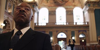 Movie still from “Heist 88” (2023), directed by Menhaj Huda – A man wearing a suit and tie in a building; Close Up shot, Low angle