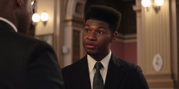 Movie still from “Heist 88” (2023), directed by Menhaj Huda – A man in a suit and tie looking at the camera; Close Up shot, Over the shoulder angle