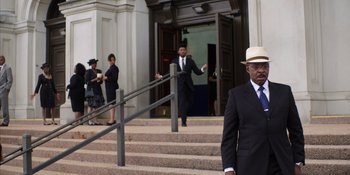 Movie still from “Heist 88” (2023), directed by Menhaj Huda – A man in a suit and hat standing in front of a building; Wide shot, Low angle