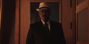 Movie still from “Heist 88” (2023), directed by Menhaj Huda – A man in a suit and a hat is standing in front of a mirror; Close Up shot, Low angle