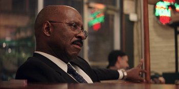 Movie still from “Heist 88” (2023), directed by Menhaj Huda – An older man sitting at a table in front of a neon sign; Close Up shot, Over the shoulder angle