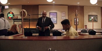 Movie still from “Heist 88” (2023), directed by Menhaj Huda – A man in a suit and tie standing next to another man in a yellow shirt; Medium shot, Low angle