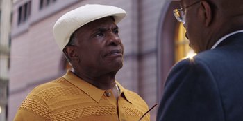Movie still from “Heist 88” (2023), directed by Menhaj Huda – An older man wearing a white hat and a yellow shirt; Close Up shot, Over the shoulder angle