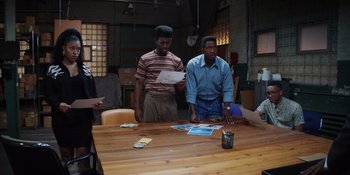 Movie still from “Heist 88” (2023), directed by Menhaj Huda – A group of people sitting around a table; Medium shot, High angle