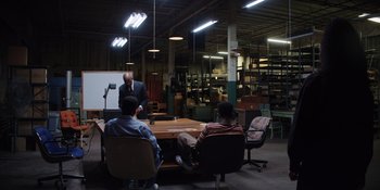 Movie still from “Heist 88” (2023), directed by Menhaj Huda – A group of people sitting around a table in a room; Wide shot, High angle