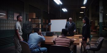 Movie still from “Heist 88” (2023), directed by Menhaj Huda – A man in a business suit is giving a presentation to a group of people; Wide shot, Over the shoulder angle