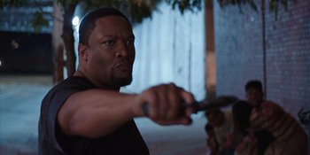 Movie still from “Heist 88” (2023), directed by Menhaj Huda – A man holding a pair of scissors in one hand and a knife in the other; Medium shot, Over the shoulder angle