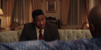 Movie still from “Heist 88” (2023), directed by Menhaj Huda – A man in a suit sitting in front of a couch; Medium shot, Over the shoulder angle