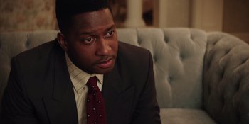 Movie still from “Heist 88” (2023), directed by Menhaj Huda – A man wearing a suit and tie sitting on a couch; Close Up shot, Over the shoulder angle