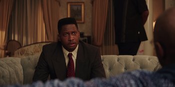 Movie still from “Heist 88” (2023), directed by Menhaj Huda – A man in a suit sitting in front of a couch; Close Up shot, Over the shoulder angle