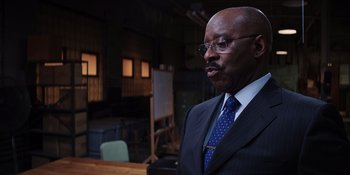 Movie still from “Heist 88” (2023), directed by Menhaj Huda – A man in a suit and tie standing in front of a painting; Close Up shot, Low angle