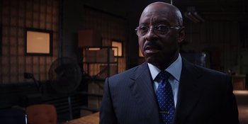 Movie still from “Heist 88” (2023), directed by Menhaj Huda – A man in a suit and tie standing in front of a fan; Close Up shot, Over the shoulder angle