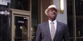 Movie still from “Heist 88” (2023), directed by Menhaj Huda – A man in a suit and a hat is standing outside; Medium shot, Low angle