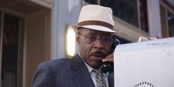 Movie still from “Heist 88” (2023), directed by Menhaj Huda – An older man in a suit and hat talking on a phone; Close Up shot, Low angle