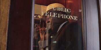 Movie still from “Heist 88” (2023), directed by Menhaj Huda – A man talking on a phone while sitting in front of a window; Extreme Close Up shot, Low angle