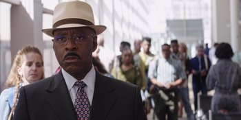 Movie still from “Heist 88” (2023), directed by Menhaj Huda – A man in a suit and a hat standing in front of a group of people; Close Up shot, Over the shoulder angle
