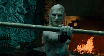 Movie still from “Hellboy II: The Golden Army” (2008), directed by Guillermo del Toro – A shirtless man holding a wooden stick in front of a fireplace; Medium shot, Over the shoulder angle