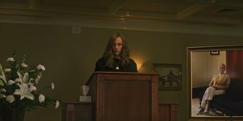 Movie still from “Hereditary” (2018), directed by Ari Aster – A person standing at a podium in a room; Medium shot, Low angle