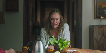 Movie still from “Hereditary” (2018), directed by Ari Aster – A woman sitting at a table with a plant in front of a tea pot; Medium shot, Over the shoulder angle