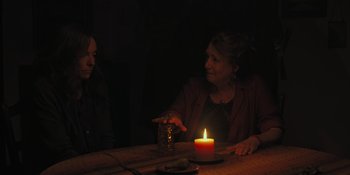 Movie still from “Hereditary” (2018), directed by Ari Aster – Two women sitting at a table in front of a lit candle; Medium shot, Over the shoulder angle