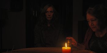 Movie still from “Hereditary” (2018), directed by Ari Aster – A woman sitting in front of a lit candle in the dark; Medium shot, Over the shoulder angle