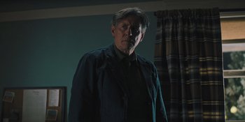 Movie still from “Hereditary” (2018), directed by Ari Aster – A man standing in a dark room with his head turned to the side; Medium shot, Low angle