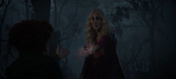 Movie still from “Hocus Pocus 2” (2022), directed by Anne Fletcher – A woman with blonde hair and a red dress is holding a glowing object in front of a forest; Medium shot, Over the shoulder angle