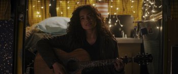 Movie still from “Holiday Harmony” (2022), directed by Shaun Paul Piccinino – A woman holding a guitar in a room with lights in the background; Medium shot, Over the shoulder angle