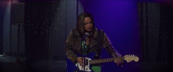 Movie still from “Holiday Harmony” (2022), directed by Shaun Paul Piccinino – A woman holding a guitar and a microphone in front of a microphone stand; Medium shot, High angle