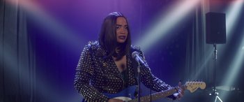 Movie still from “Holiday Harmony” (2022), directed by Shaun Paul Piccinino – A woman in a leather jacket holding a blue guitar; Medium shot, Over the shoulder angle
