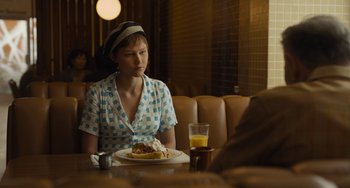 Movie still from “Hollywood Stargirl” (2022), directed by Julia Hart – A woman sitting in front of a plate of food on top of a table; Medium shot, Over the shoulder angle