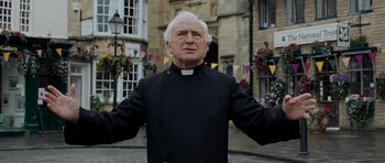 Movie still from “Hot Fuzz” (2007), directed by Edgar Wright – An older man wearing a priest's outfit in front of a building; Medium shot, Low angle
