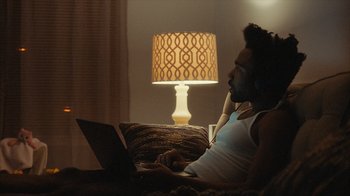 Movie still from “Atlanta” (2016), created by Donald Glover – A man sitting on a couch using a laptop computer; Medium shot, Over the shoulder angle