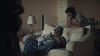 Movie still from “Atlanta” (2016), created by Donald Glover – A man sitting on top of a couch while another man looks at his phone; Medium shot, Over the shoulder angle