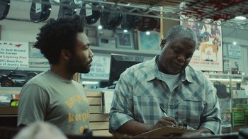 Movie still from “Atlanta” (2016), created by Donald Glover – Two men are sitting at a table and one is writing on a piece of paper; Medium shot, Over the shoulder angle