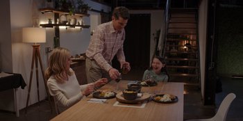 Movie still from “I Believe in Santa” (2022), directed by Alex Ranarivelo – A man standing at a dinner table with two women and a child; Medium shot, High angle