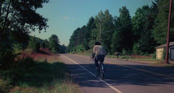 Movie still from “I Spit on Your Grave” (1978), directed by Meir Zarchi – A man riding a bike down the middle of the road; Extreme Wide shot, High angle