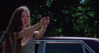 Movie still from “I Spit on Your Grave” (1978), directed by Meir Zarchi – A woman taking a picture with her cell phone while standing in front of a car; Medium shot, Low angle