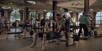 Movie still from “I Want You Back” (2022), directed by Jason Orley – A man lifting a barbell in a gym while another man watches; Wide shot, Over the shoulder angle
