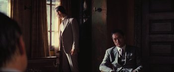 Movie still from “Inception” (2010), directed by Christopher Nolan – A man in a suit standing next to another man in a suit sitting in front of a window; Medium shot, Over the shoulder angle