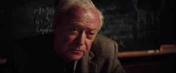 Movie still from “Inception” (2010), directed by Christopher Nolan – An older man in a suit is sitting in front of a chalkboard; Close Up shot, Over the shoulder angle