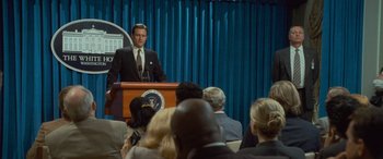 Movie still from “Independence Day” (1996), directed by Roland Emmerich – A man is speaking at a podium in front of a group of people; Medium shot, Over the shoulder angle