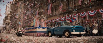 Movie still from “Indiana Jones and the Dial of Destiny” (2023), directed by James Mangold – A truck driving down a street past a parade float; Extreme Wide shot, High angle