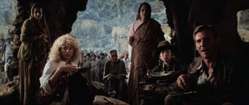Movie still from “Indiana Jones and the Temple of Doom” (1984), directed by Steven Spielberg – A group of people sitting in a cave with a woman standing in front of them; Medium shot, Low angle