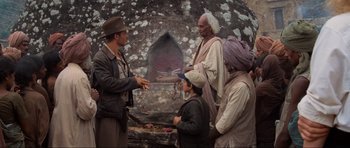 Movie still from “Indiana Jones and the Temple of Doom” (1984), directed by Steven Spielberg – A man and two women standing next to an older man; Medium shot, Over the shoulder angle