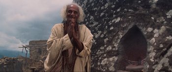 Movie still from “Indiana Jones and the Temple of Doom” (1984), directed by Steven Spielberg – An old man with a long white beard is praying in front of a rock wall; Medium shot, Low angle