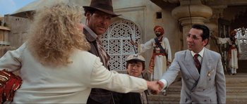 Movie still from “Indiana Jones and the Temple of Doom” (1984), directed by Steven Spielberg – A group of people standing next to each other near a building; Medium shot, Over the shoulder angle